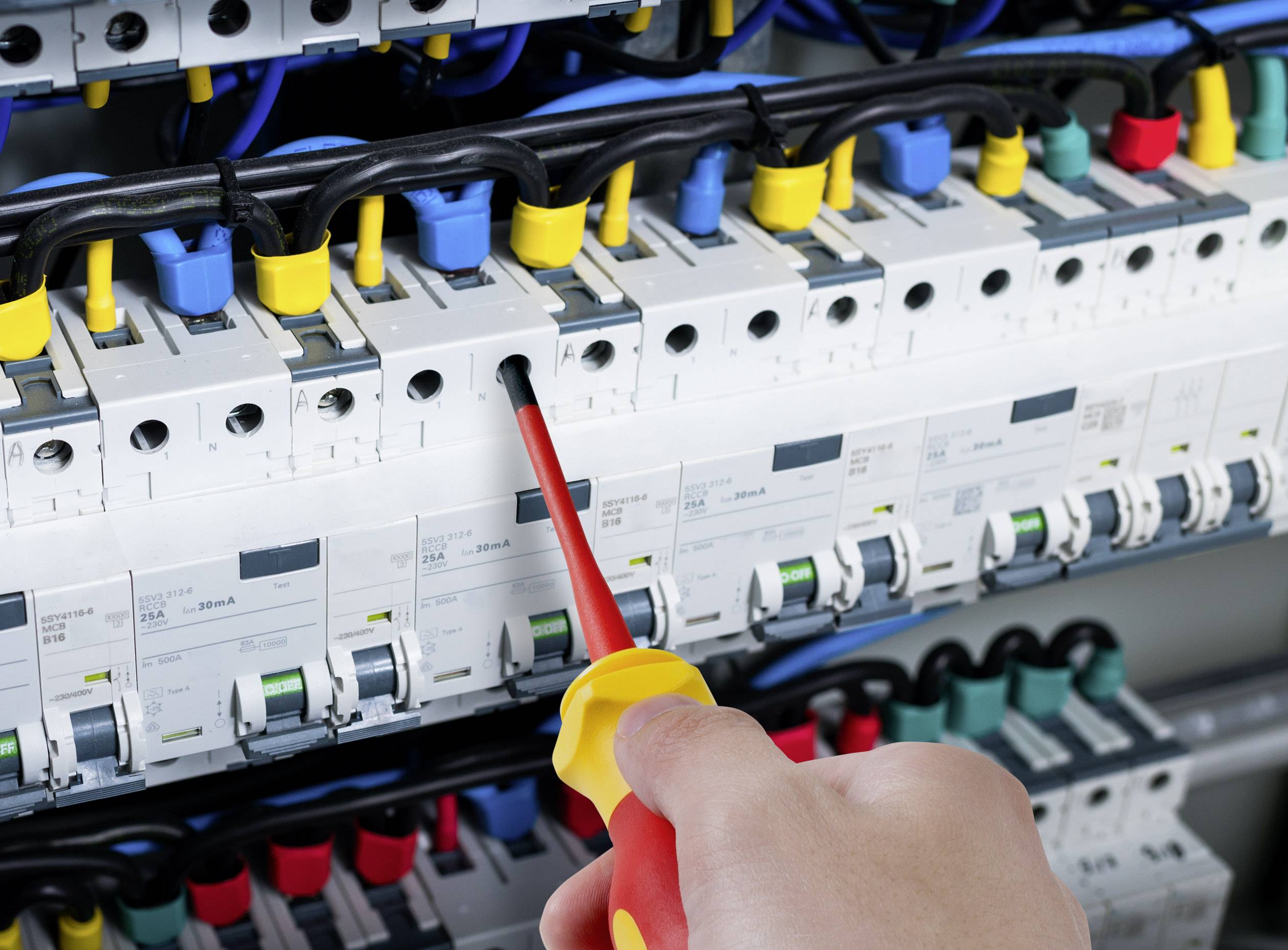 A hand holds a screwdriver and is working on a fuse box with switches and colourful wires.