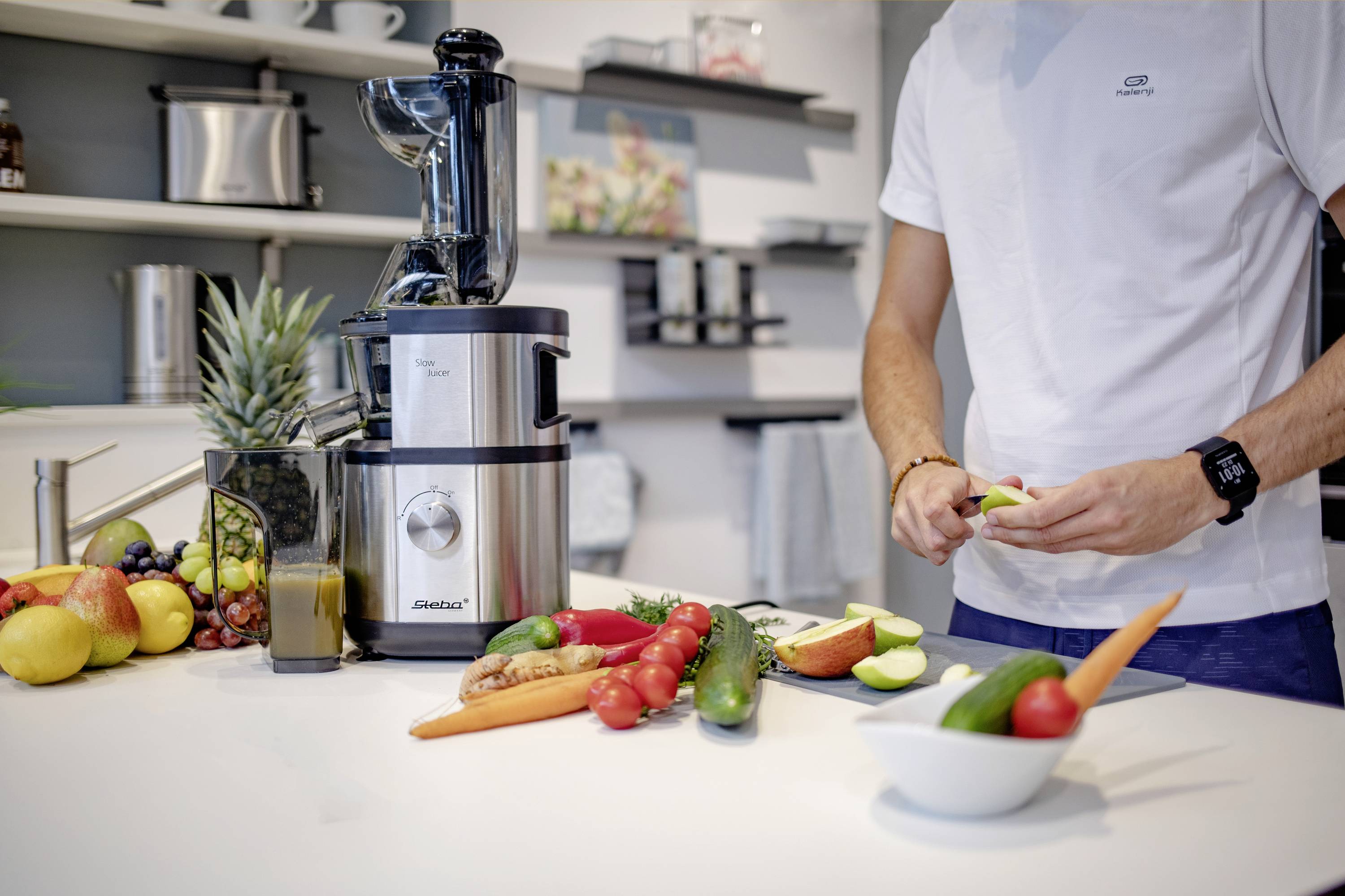 A person is preparing fruit and vegetables next to a juicer in a modern kitchen, surrounded by various fruits.