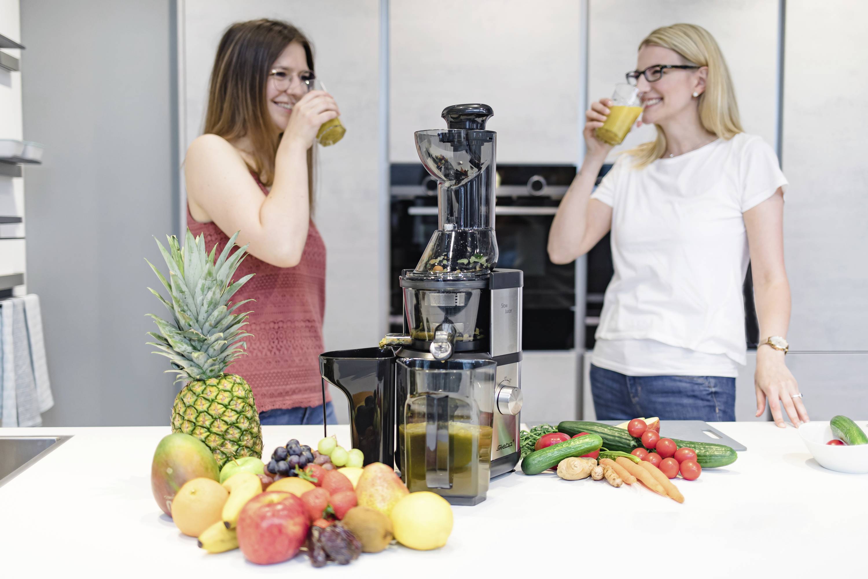 Two women are drinking juice in a modern kitchen. On the worktop, there is a juicer surrounded by fruits and vegetables such as pineapple and tomatoes.
