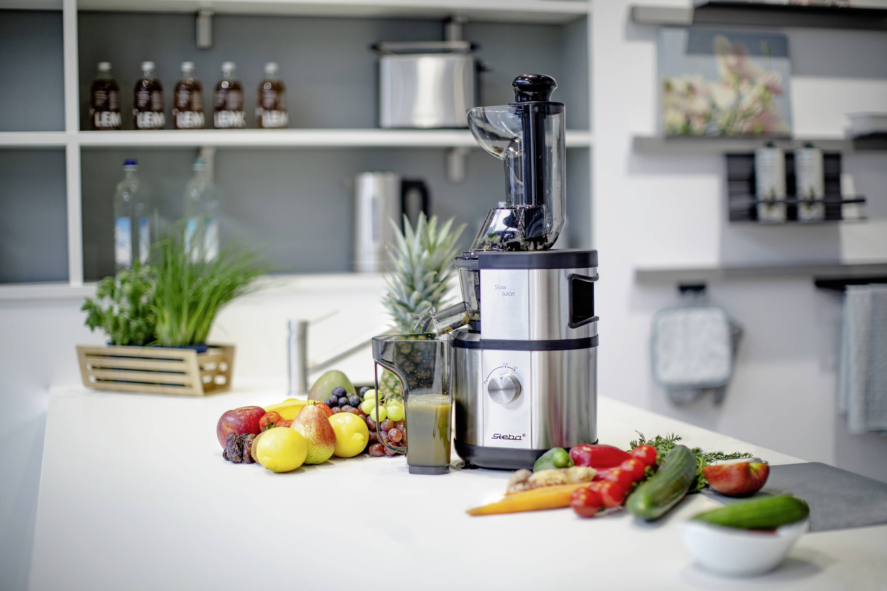 Juicer on a kitchen worktop with various fruits and vegetables such as apples, carrots and pineapple. Kitchen utensils in the background.