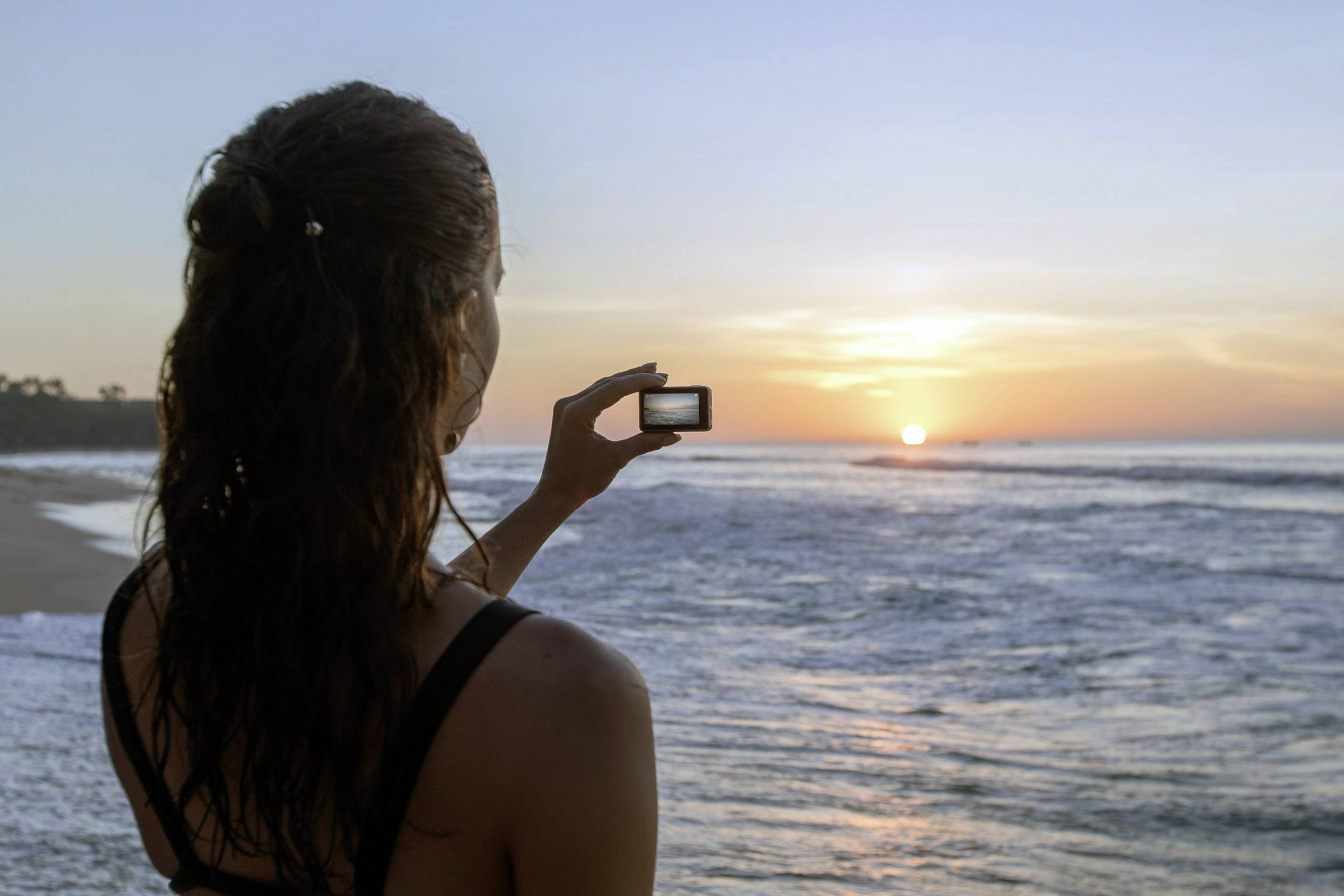 A woman on the beach photographs a sunrise over the sea. In the foreground, the glistening water and the clear morning sun are visible.