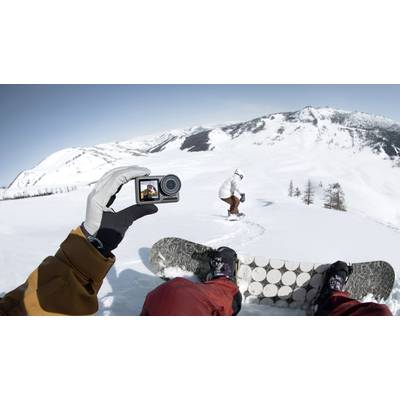 A person is snowboarding in snow-covered mountains, while someone films the scene from a first-person perspective using a small camera.