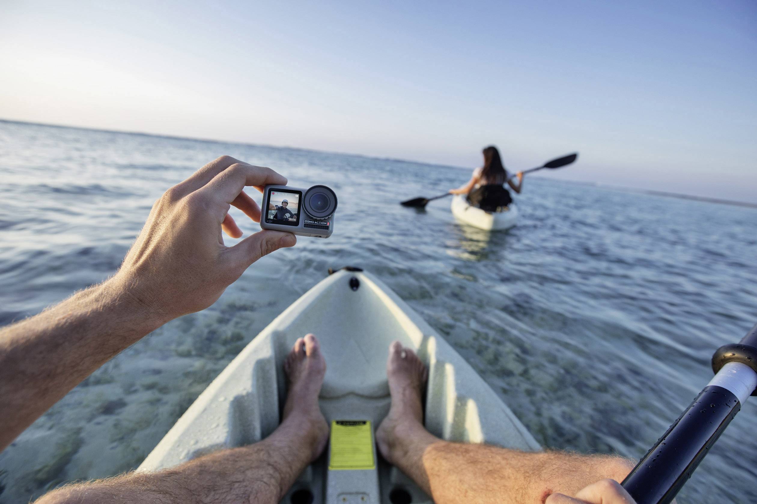 A person photographs from a kayak perspective, capturing another person paddling ahead. Vast ocean, clear sky in the background.