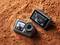 Two action cameras on red earth. The cameras display images of motocross riders in action on their screens.