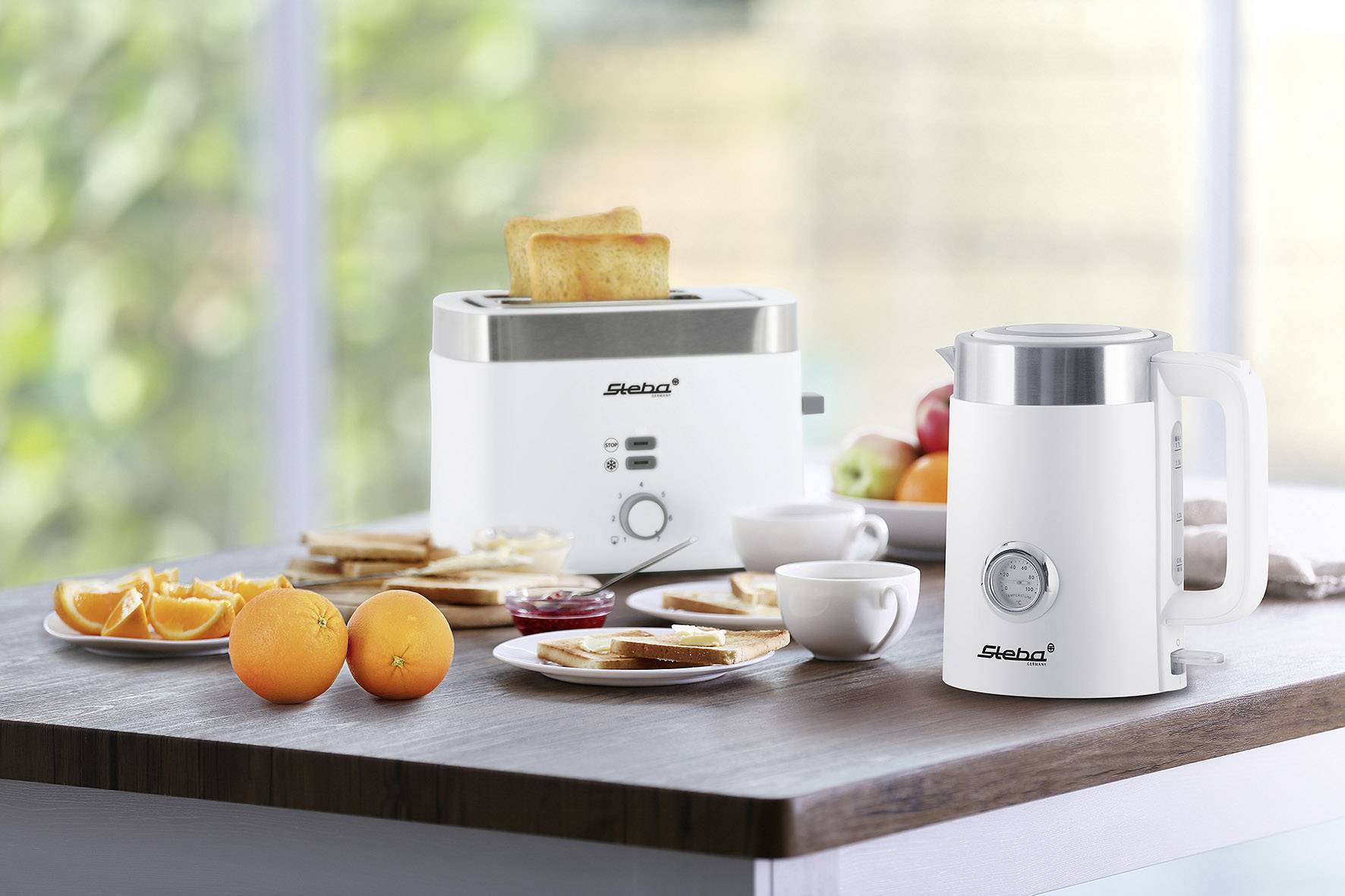 A toaster with toast slices and a kettle are standing on a table. Next to them are oranges, jam, and a cup.