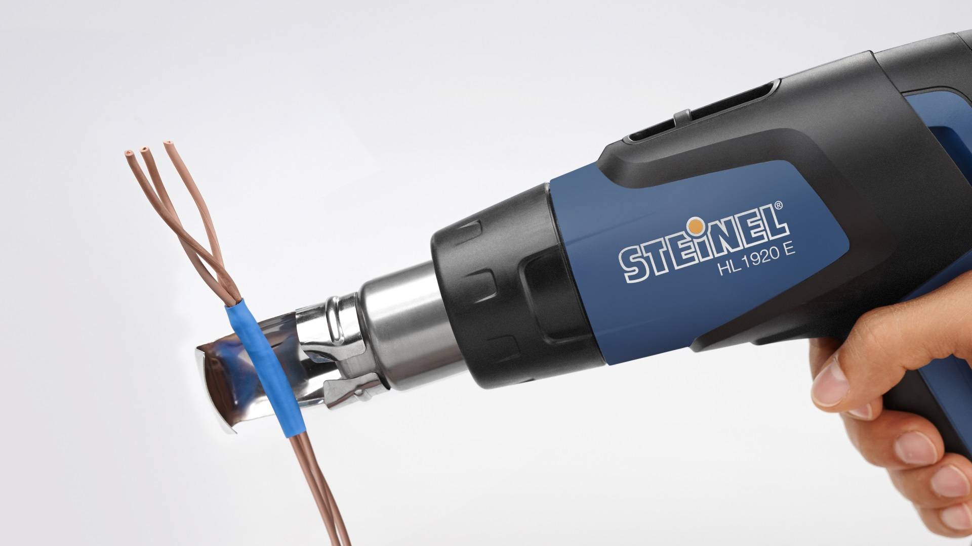 A hand is holding a Steinel HL 1920 E heat gun, which is directed at electrical cables to shrink insulation.