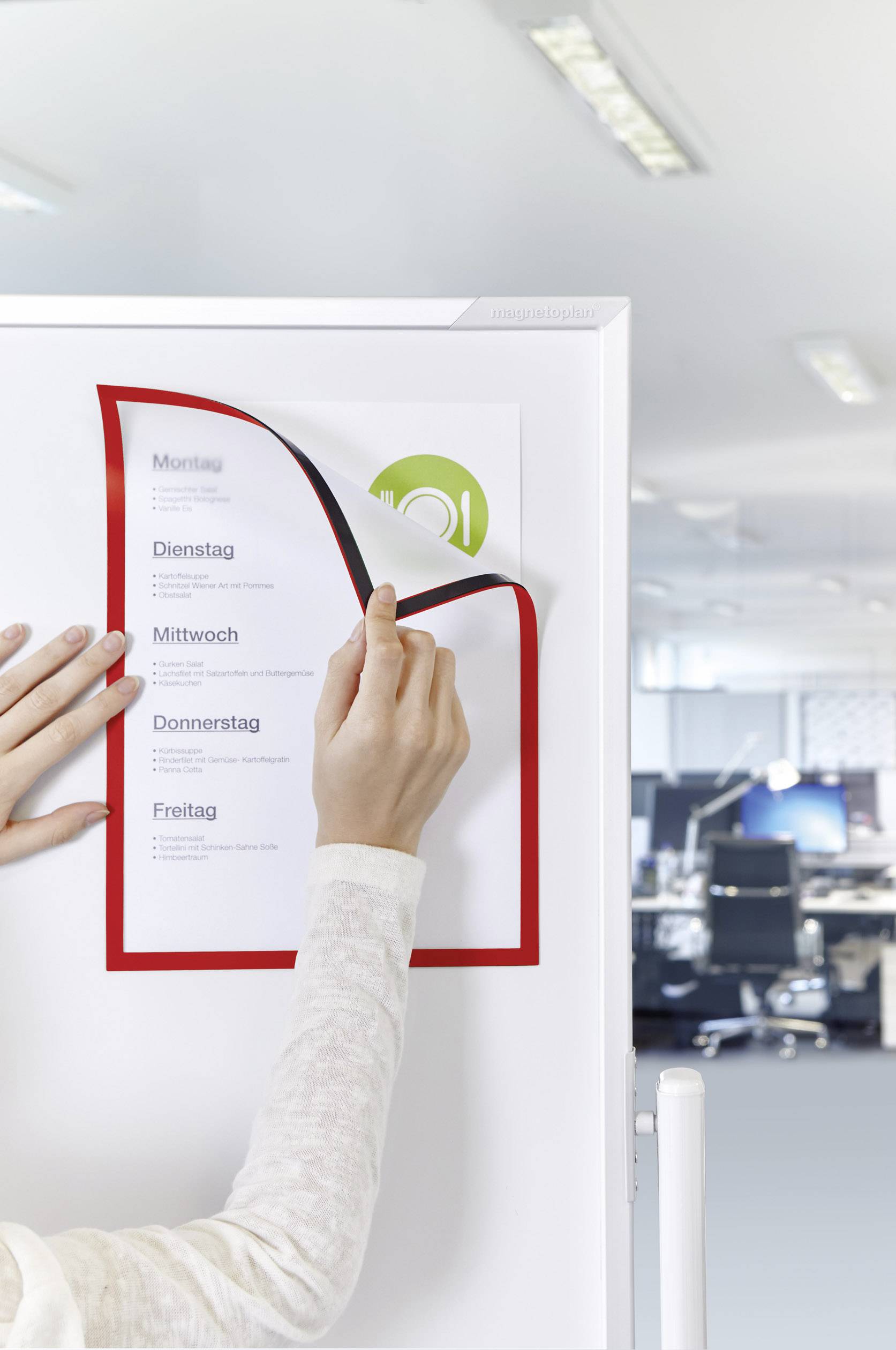 A person is pointing to a magnetic weekly planner board on a fridge in an office. The board displays the weekdays from Monday to Friday.