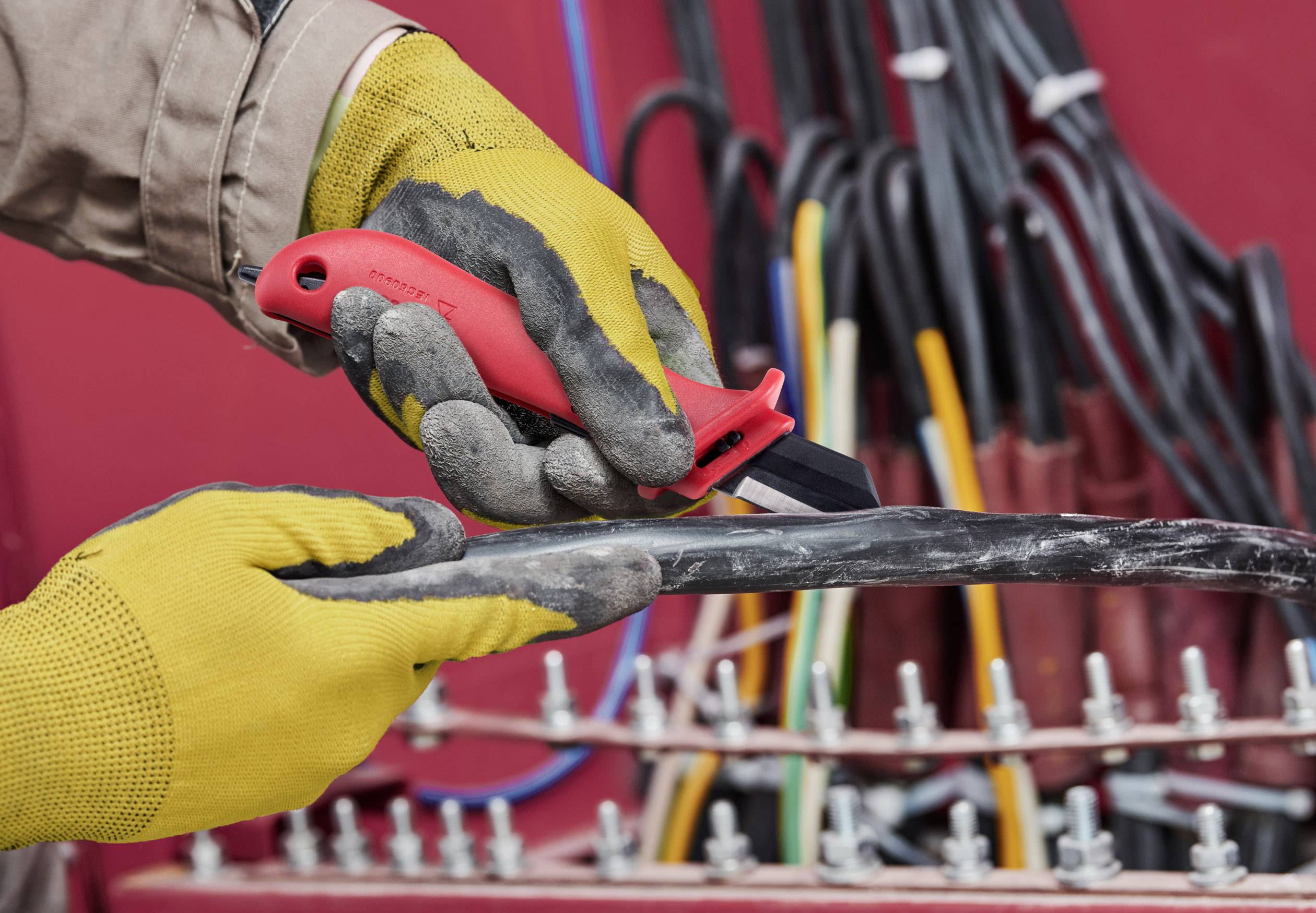 A person wearing work gloves is stripping a thick electrical cable with a red utility knife, with additional cables visible in the background.