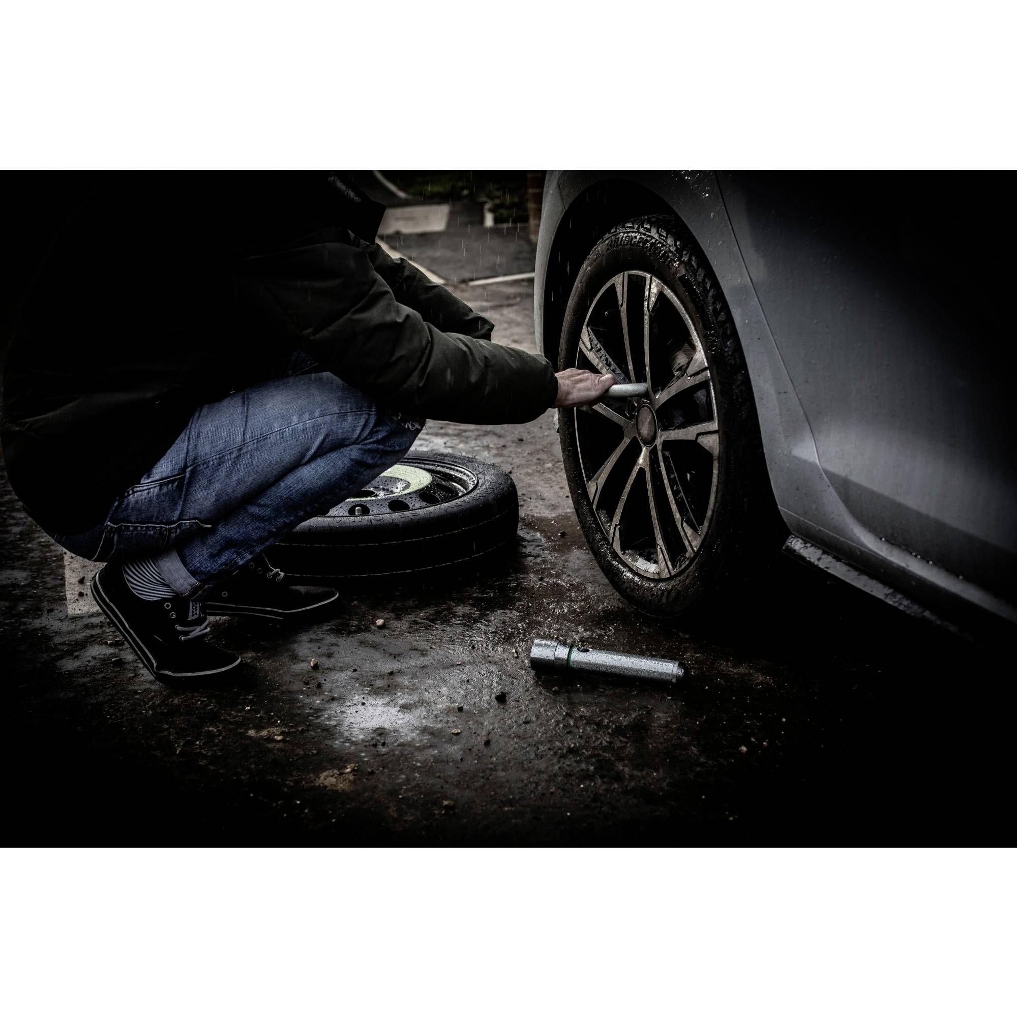 A person is changing a car tyre in poor light. A spare tyre and tools are visible on the ground.