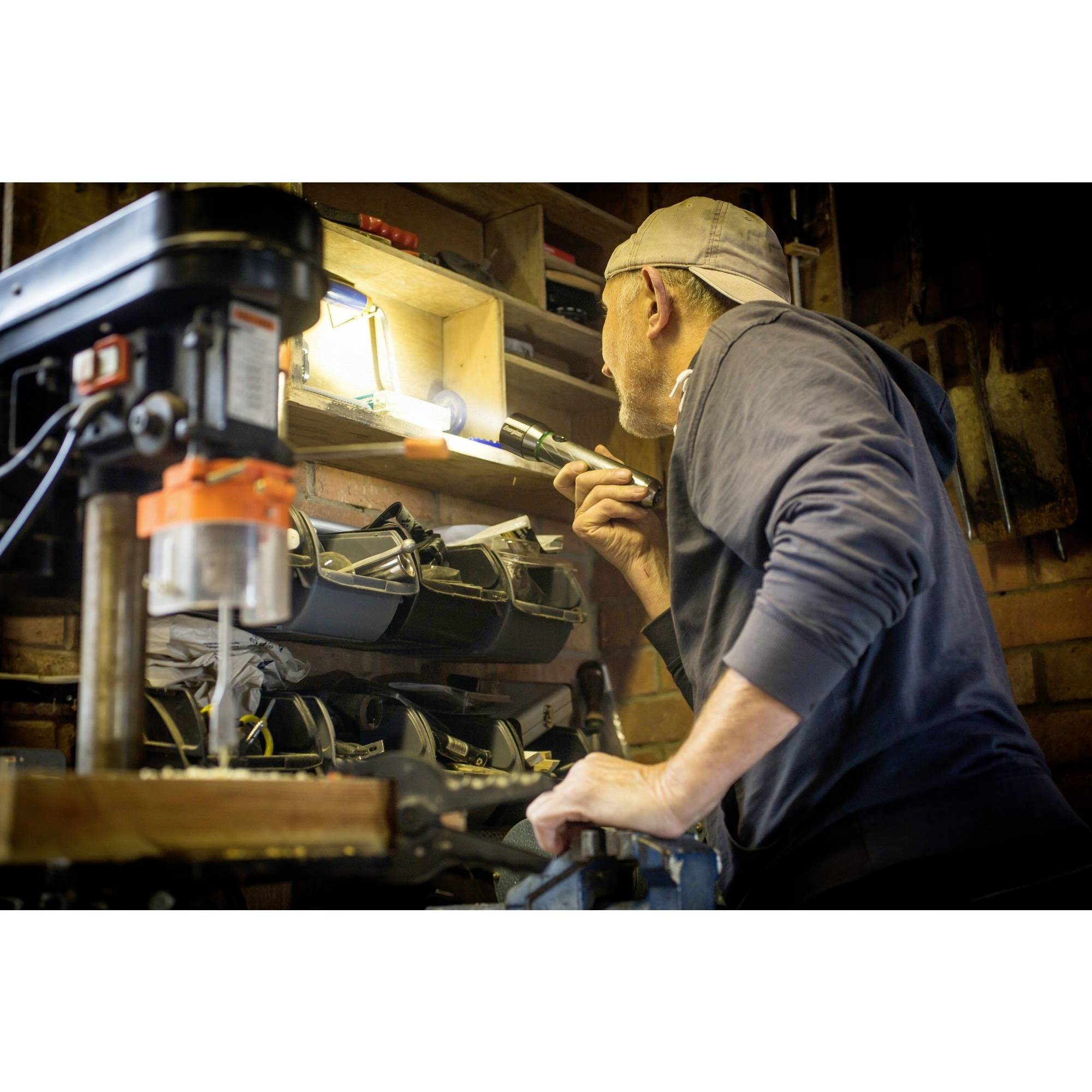 A man in a workshop is using a torch to search through items on a shelf. Tools are visible in the foreground.