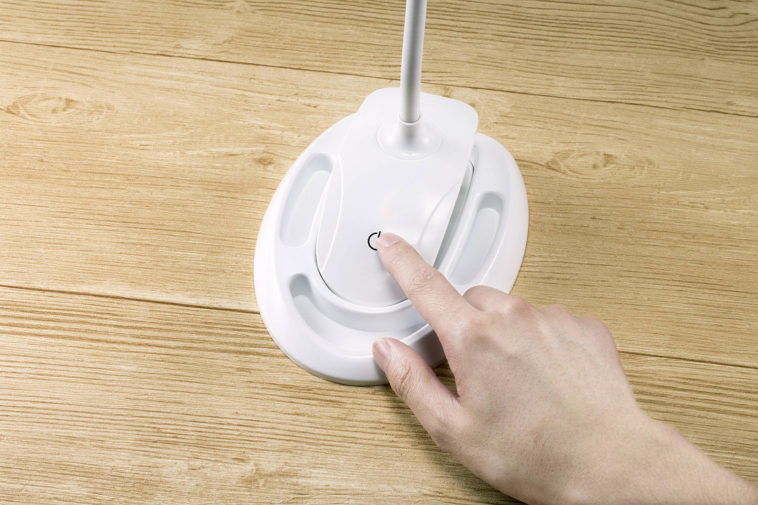 A hand presses the power button on a white stand on a wooden surface.