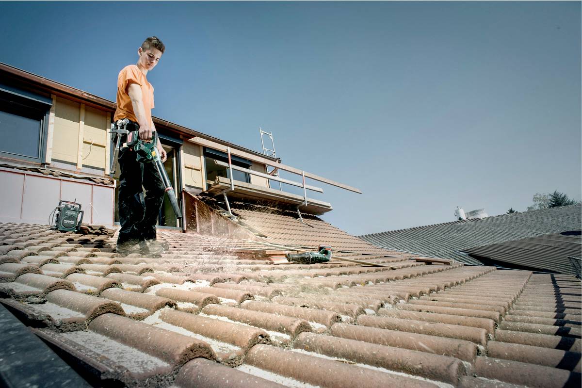 A person is cleaning a sloping tiled roof of a house with a pressure washer, surrounded by a clear sky and other houses.
