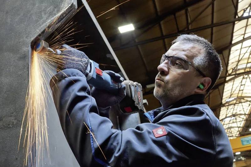 A man wearing safety gear grinds a metal surface, causing bright sparks to fly. He is focused and surrounded by industrial equipment.