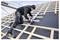 A worker in safety gear kneels on a roof's wooden framework, using a circular saw to cut a horizontal beam. Scaffolding visible in the background.