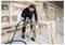 A person in a workshop uses a power tool to cut a wooden board on a sawhorse, surrounded by unfinished wooden framing and drywall.