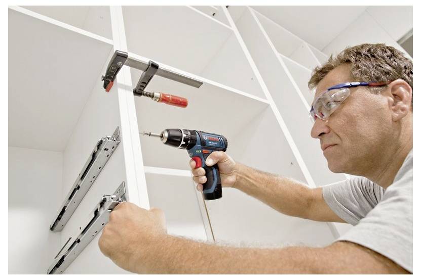 A man wearing safety glasses uses a power drill to install cabinet fixtures in a white shelving unit.