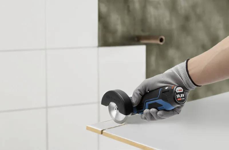 A person wearing a glove uses a compact power tool to cut a tile. The tool is black and blue, and the setting appears to be a work area.