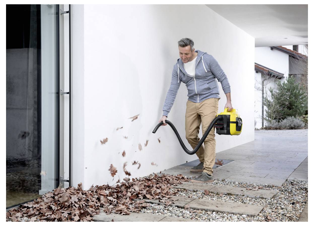 A person using a leaf blower on a patio to clear fallen leaves, dressed casually with a focused expression, near a modern building.