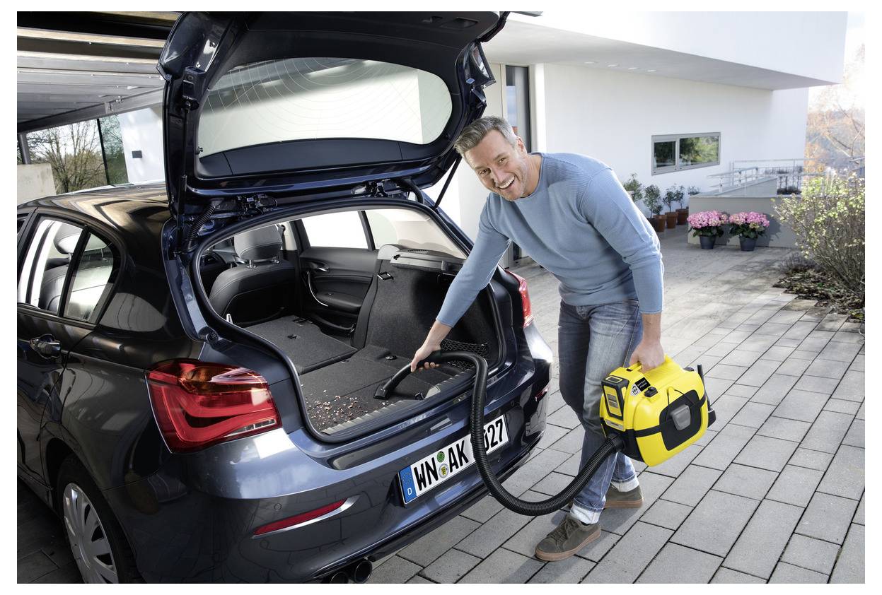 A man uses a yellow portable vacuum to clean the trunk of a blue car parked in a driveway.