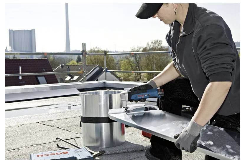 'Person using a Bosch power tool to cut metal on a rooftop, surrounded by safety equipment and distant buildings in the background.'