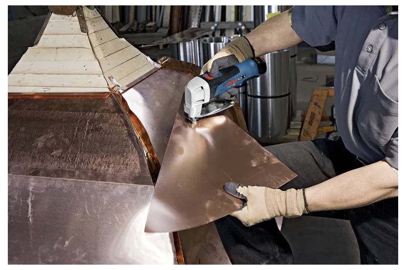 A person uses an electric tool to cut a sheet of copper metal, wearing gloves for safety. The setting appears to be an industrial workshop.
