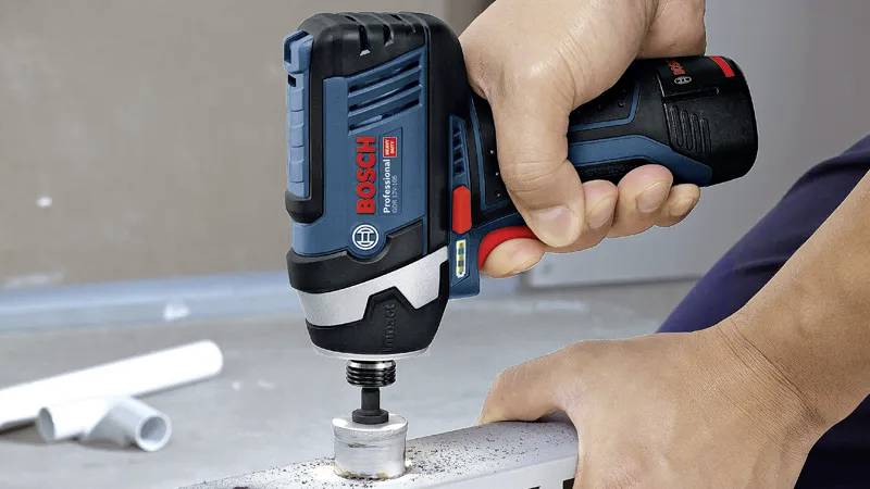 A person uses a Bosch cordless impact driver on a metal surface, screwing a bolt. The tool is black and blue, and the environment is industrial.