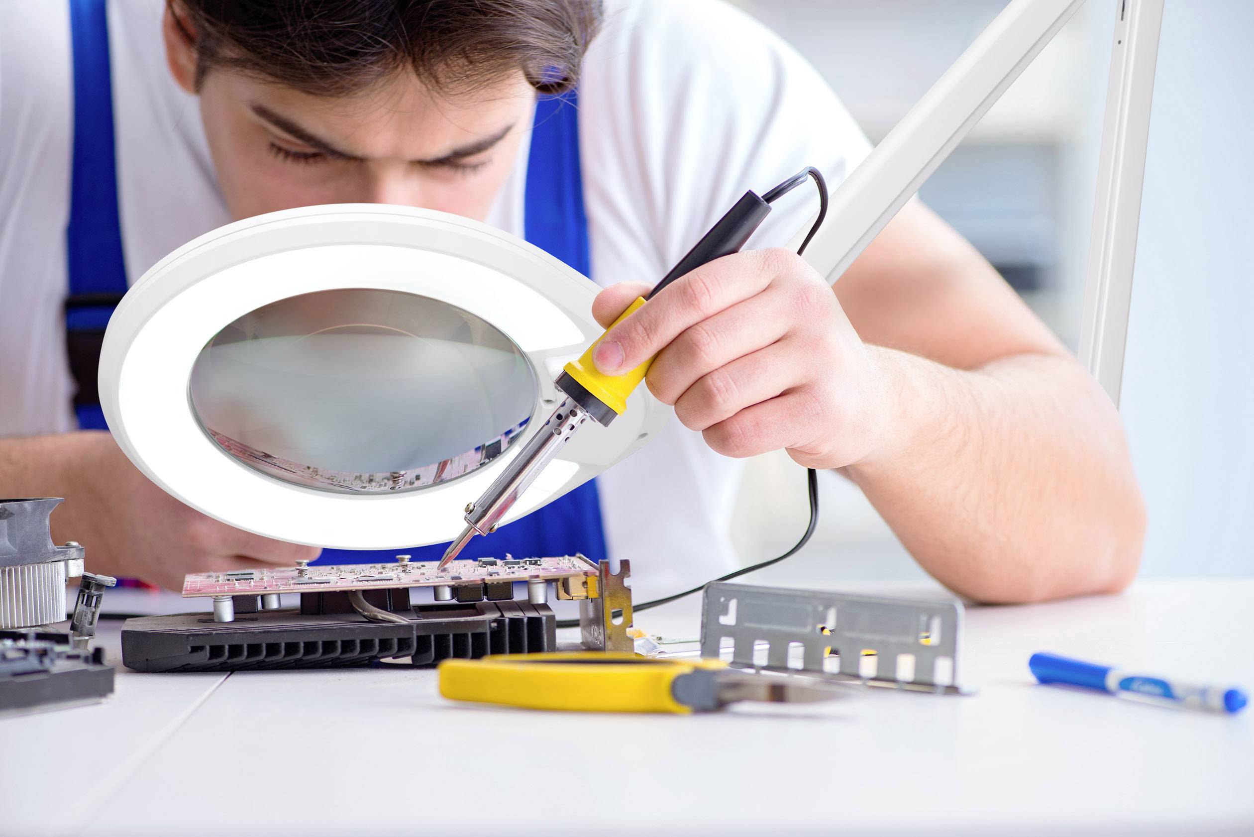 A technician solders carefully on a circuit board under a magnifying glass, surrounded by tools.