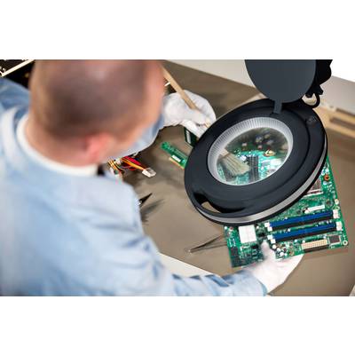 A technician is cleaning an exposed circuit board with a brush under an illuminated magnifying glass on a table.