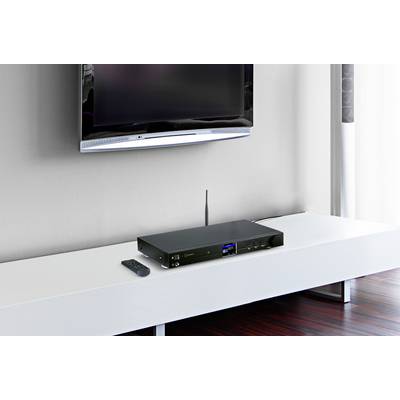 A modern black receiver sits on a white TV console. Next to it is a remote control. In the background, a flatscreen television is mounted on the wall.