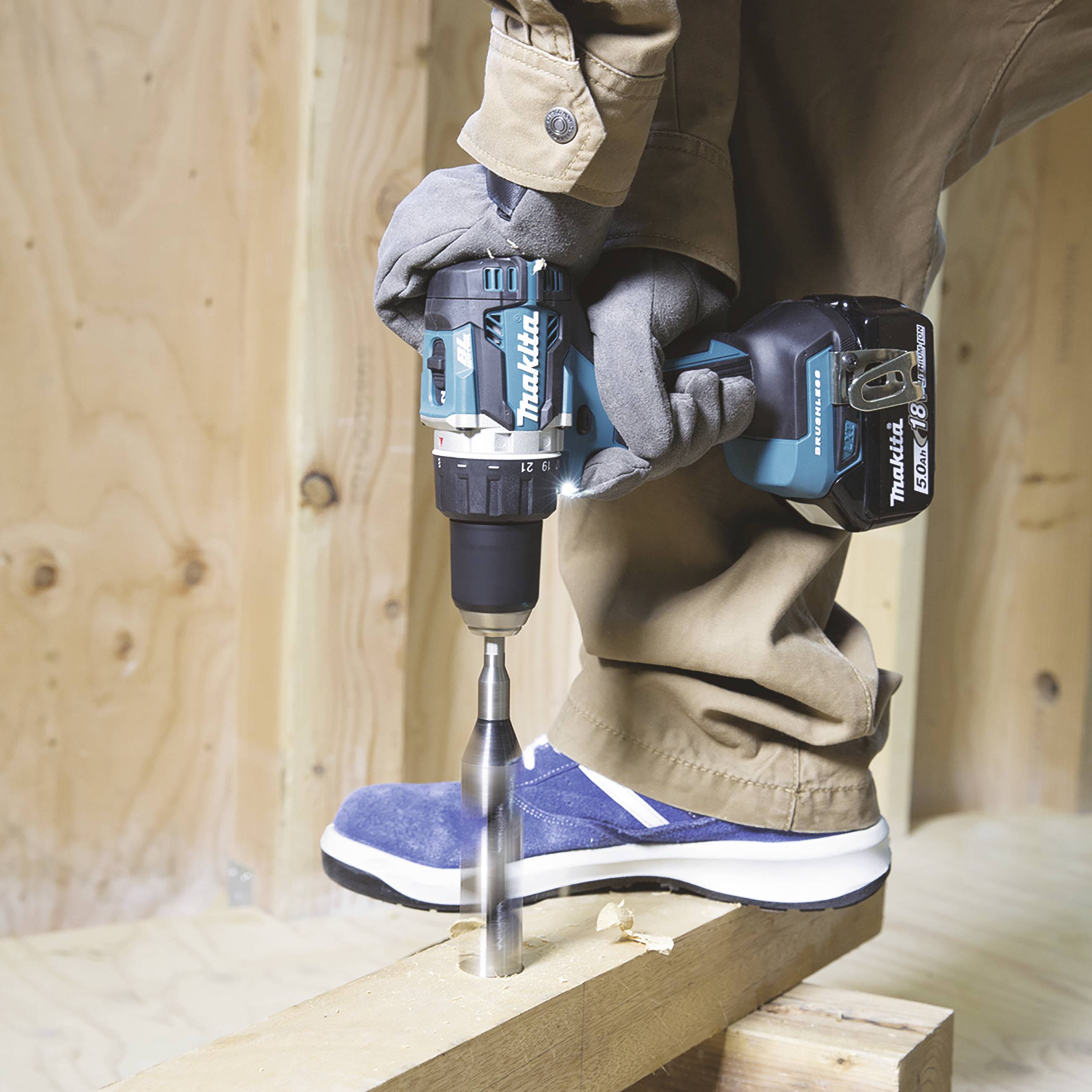 A person in work attire is using a cordless drill to bore a hole into a wooden board. The surrounding environment is a construction site.