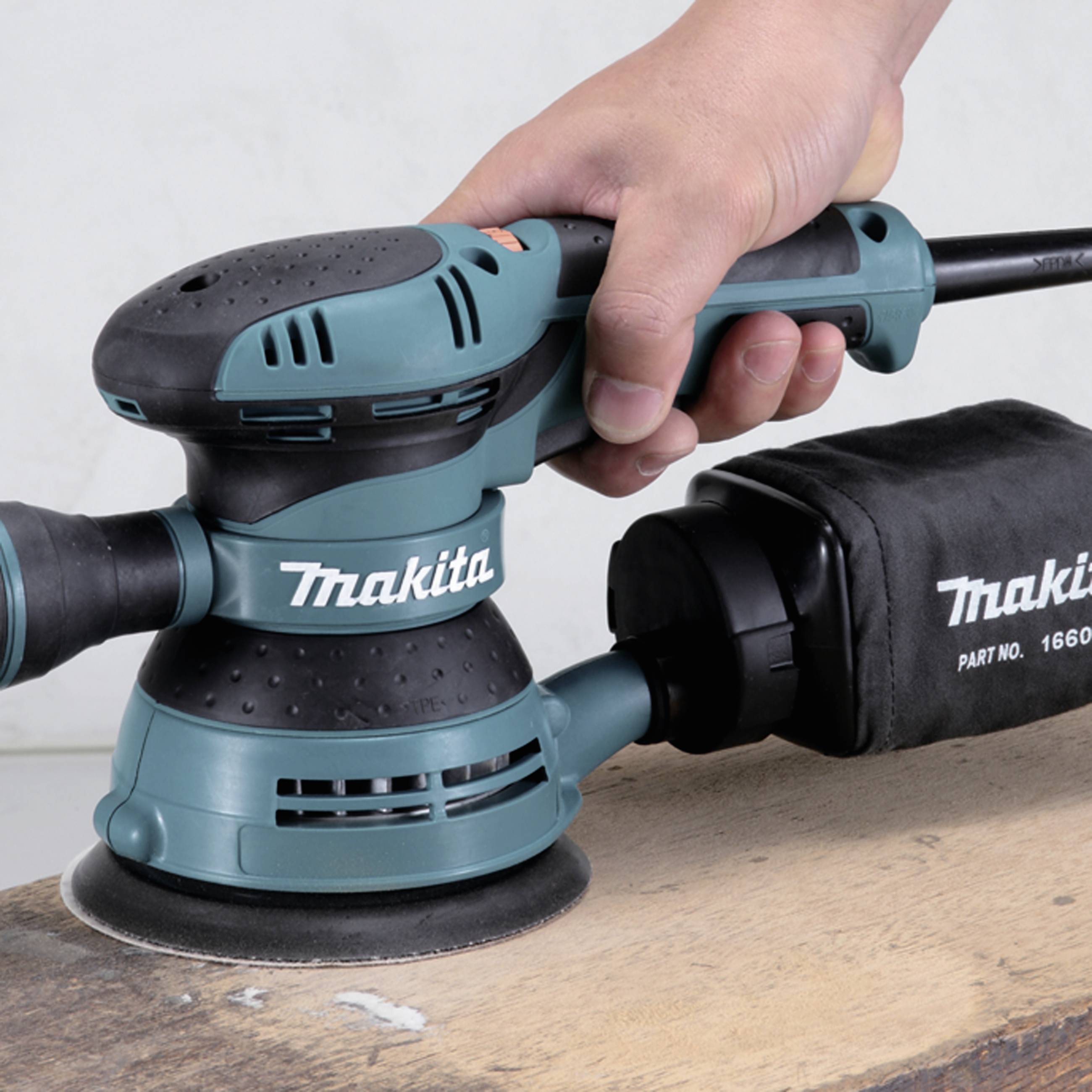 A hand is holding a green, electric orbital sander by Makita on a wooden surface, ready for use.