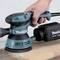 A hand is holding a green, electric orbital sander by Makita on a wooden surface, ready for use.