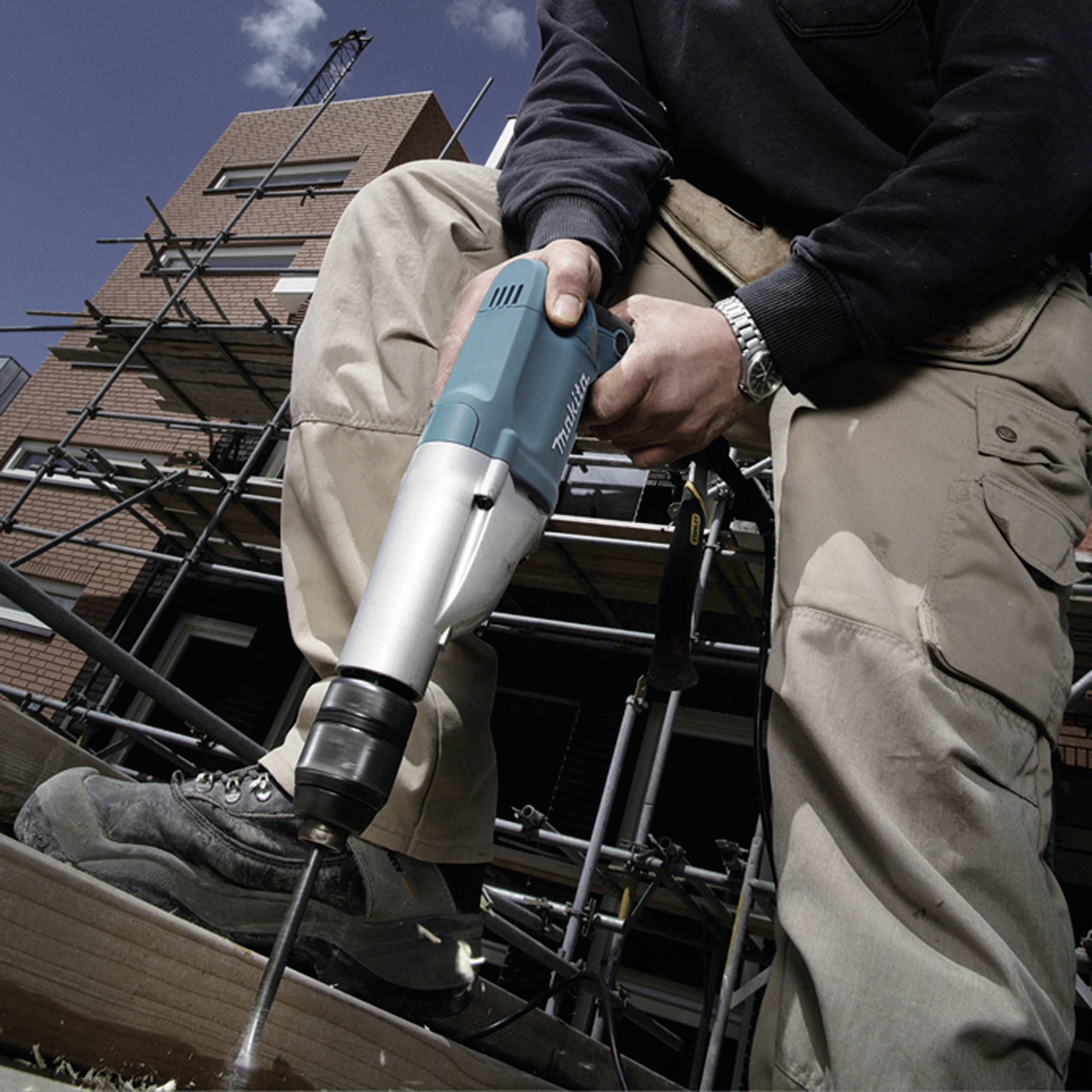 A person is using an electric drill on a construction site, focusing on drilling into a wooden board. A scaffolding and a house are visible in the background.