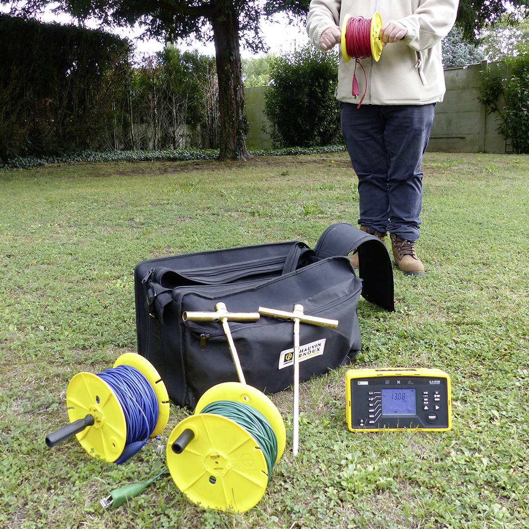 A person is holding a cable reel, with additional cable reels, a device, and a bag on the ground nearby. Trees can be seen in the background.