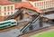 Model railway depicts a station with a train, pedestrian bridge, and figures. The main building has red roofs. People are moving on the bridge and the tracks.