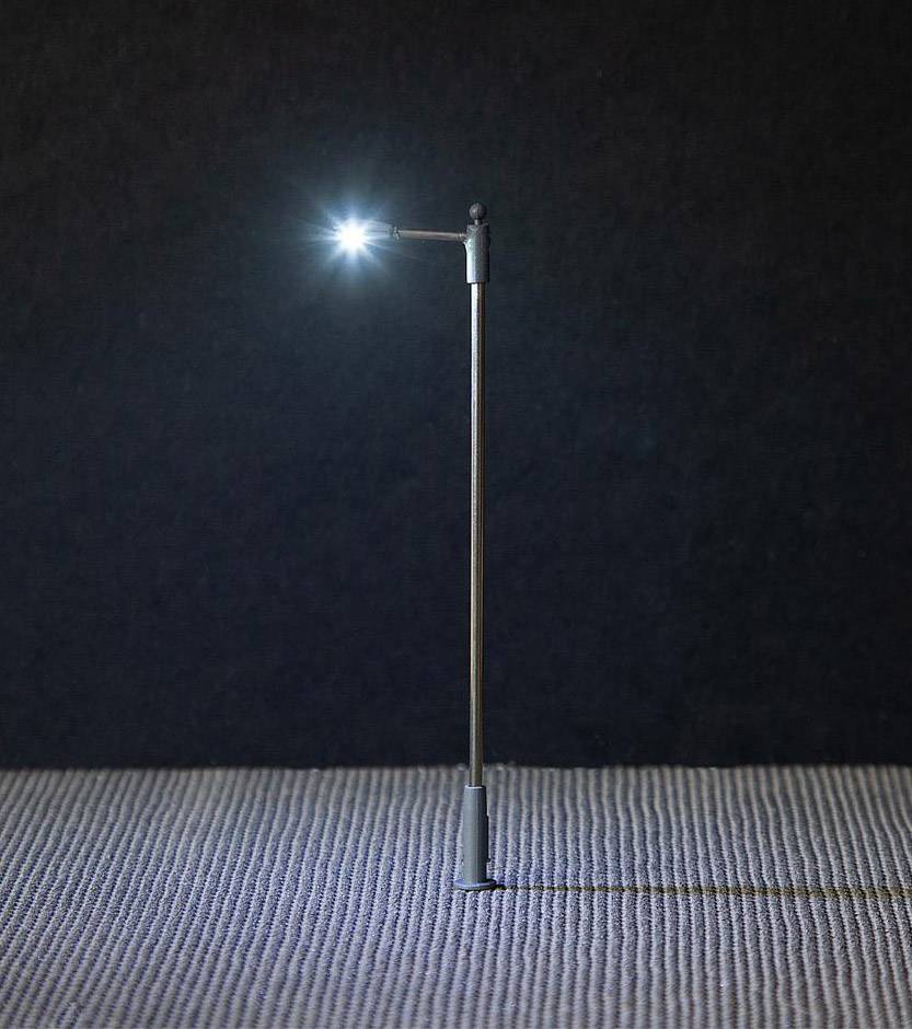A single, minimalist street lamp illuminates against a dark background, casting light on the textured ground beneath.