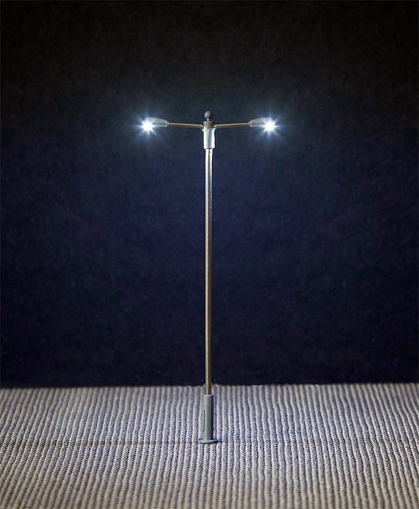 A miniature street lamp with two illuminating lamps stands on textured ground against a dark background.