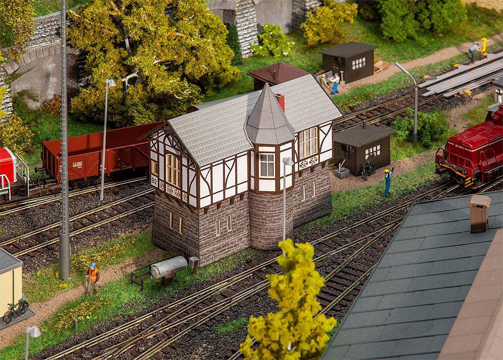 'Miniature railway scene with signal box, surrounded by tracks and model trains. Figures stand beside the rails, surrounded by trees.'