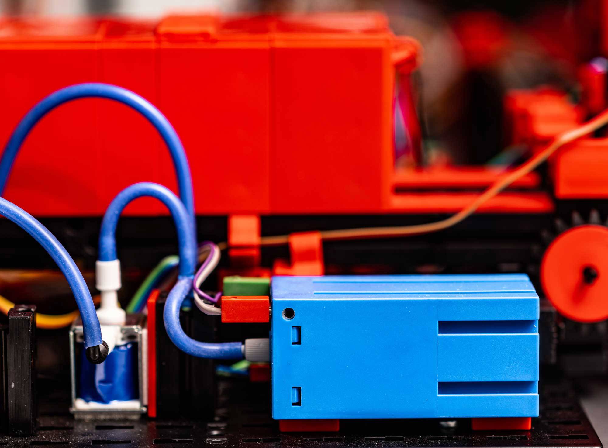 A detail of a toy train with red and blue building blocks, connected by colourful cables. Background blurred.
