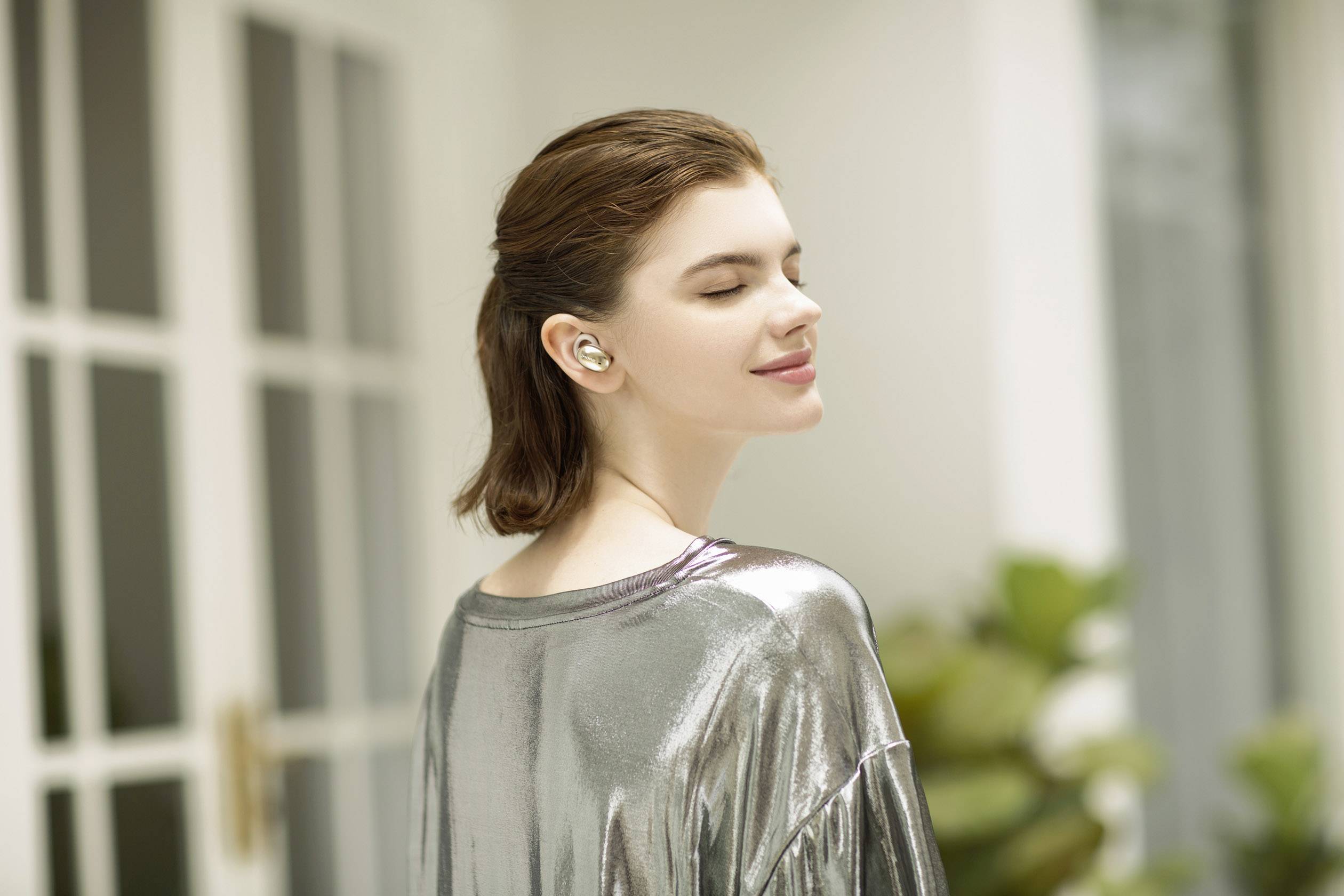 A woman with closed eyes is wearing wireless earbuds and a shiny top. She stands relaxed in a bright room.