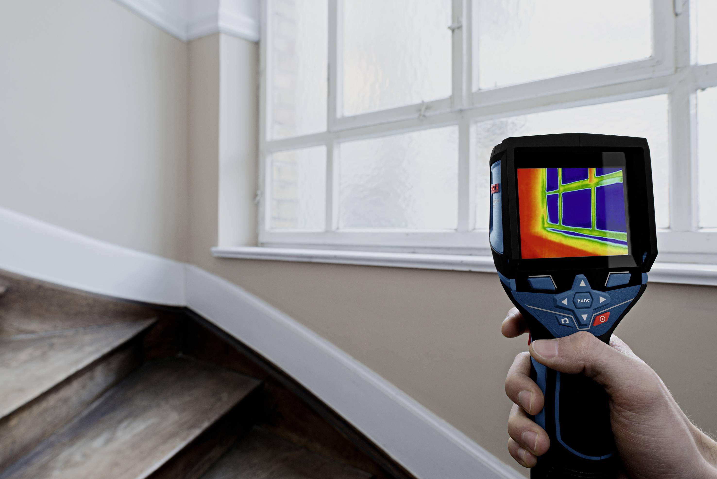 A hand is holding a thermal imaging camera, which displays a window and temperature variations in the room.