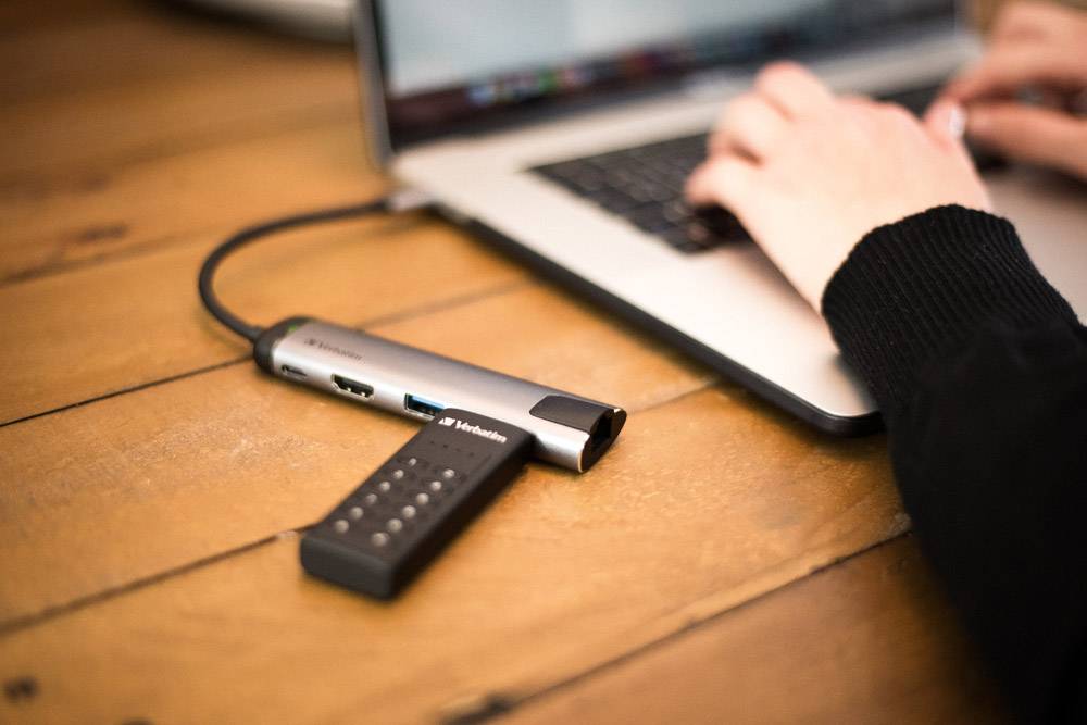 A person is typing on a laptop. A USB hub with multiple ports is connected and lies on a wooden table.