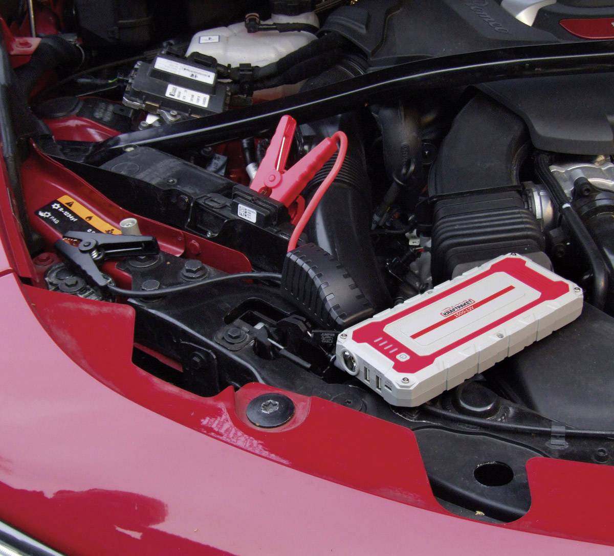 A car battery in the engine compartment is being jump-started with a portable jump starter. Red and black cables are connected.