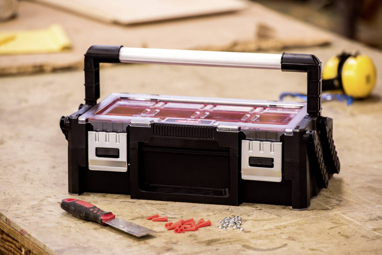 A black toolbox with a transparent lid sits on a workbench. Beside it are a knife, wall plugs, and screws.