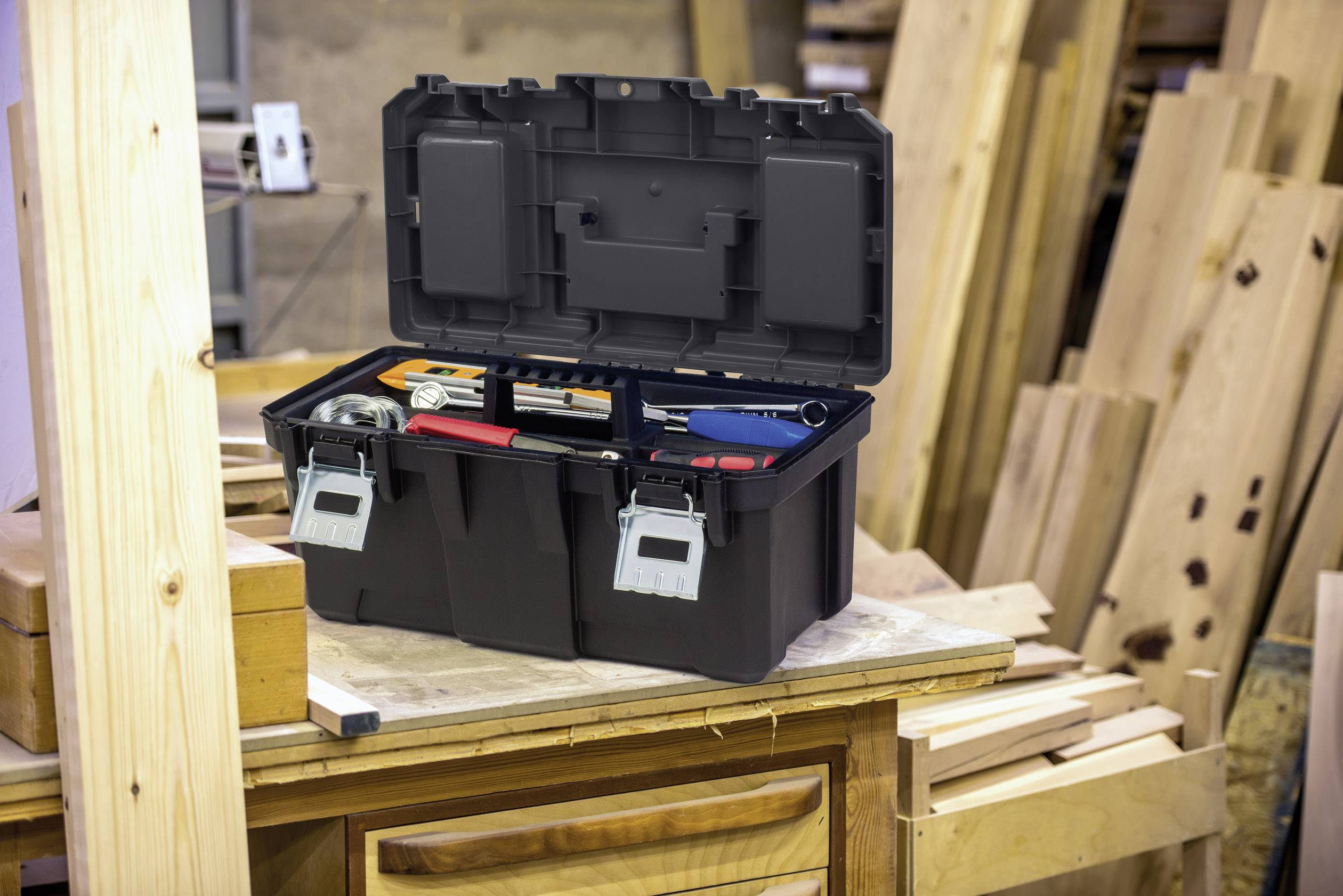 An open toolbox with various tools sits on a wooden workbench, surrounded by wooden boards in a workshop.
