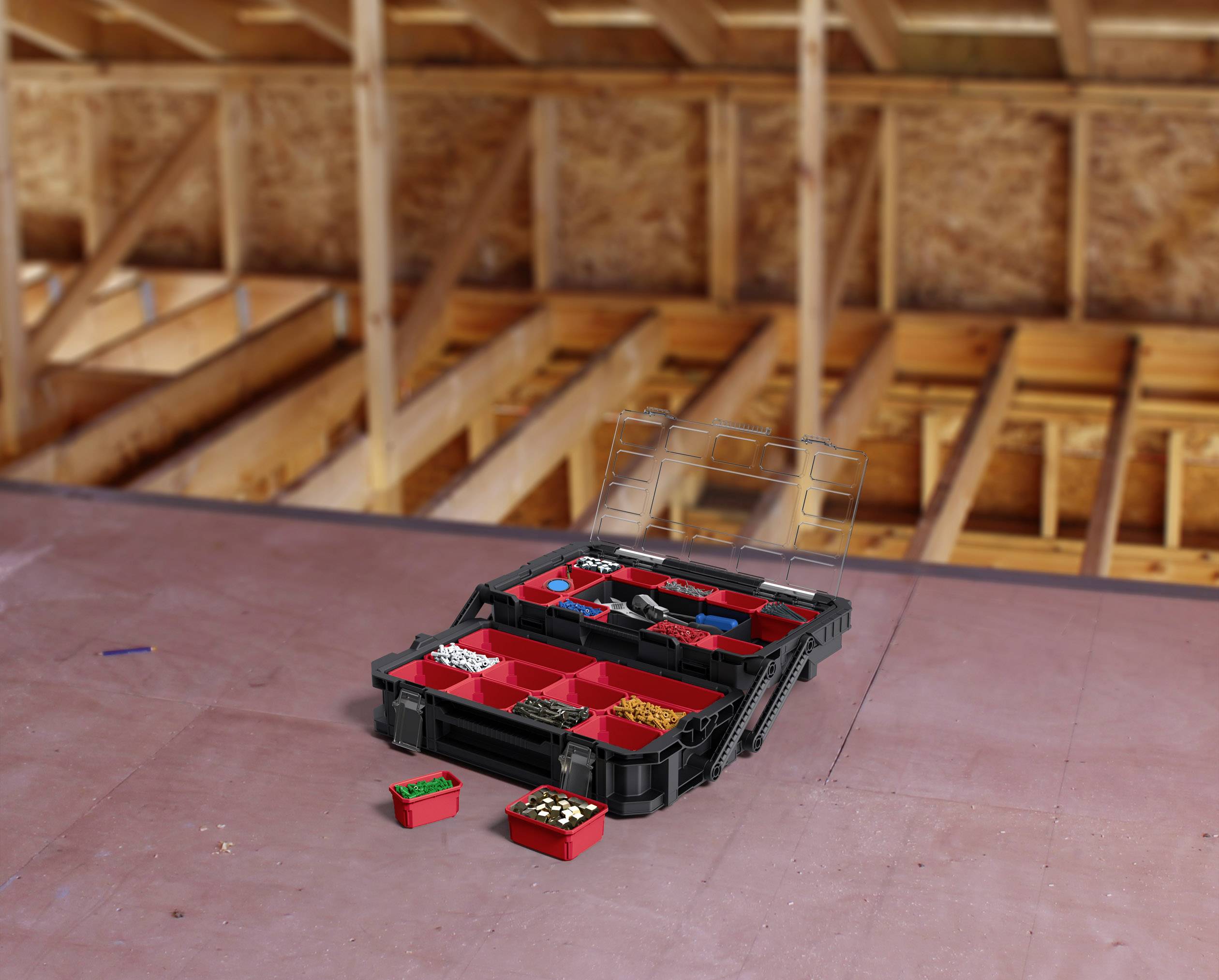 Black toolbox with red drawers full of screws and nails, placed on the floor of an attic under construction.