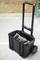 Black wheeled suitcase with extendable handle, containing an electric tool. Background of wooden panelling and concrete floor.