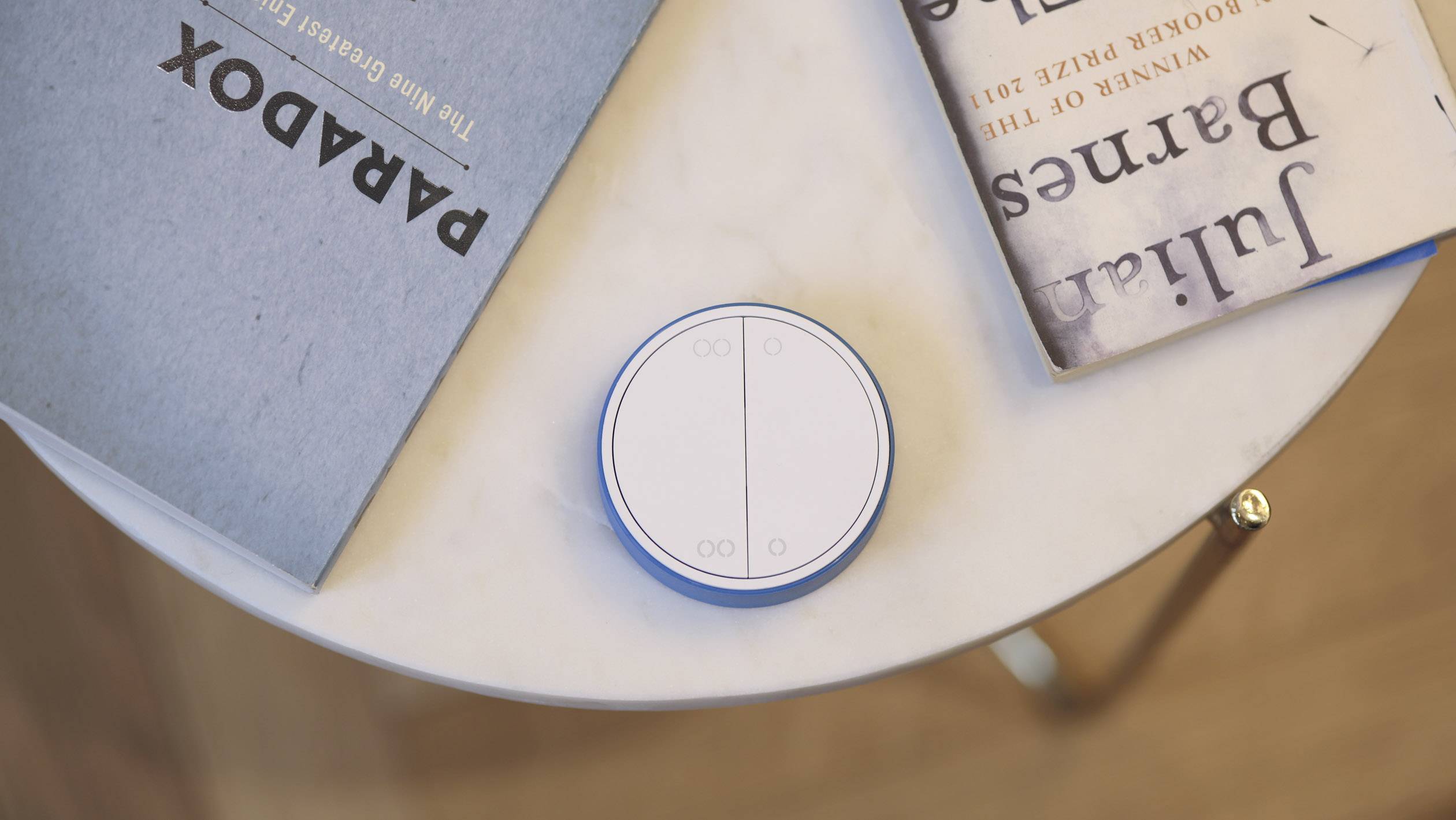 On a marble table, a round, white smart home controller lies beside two books.