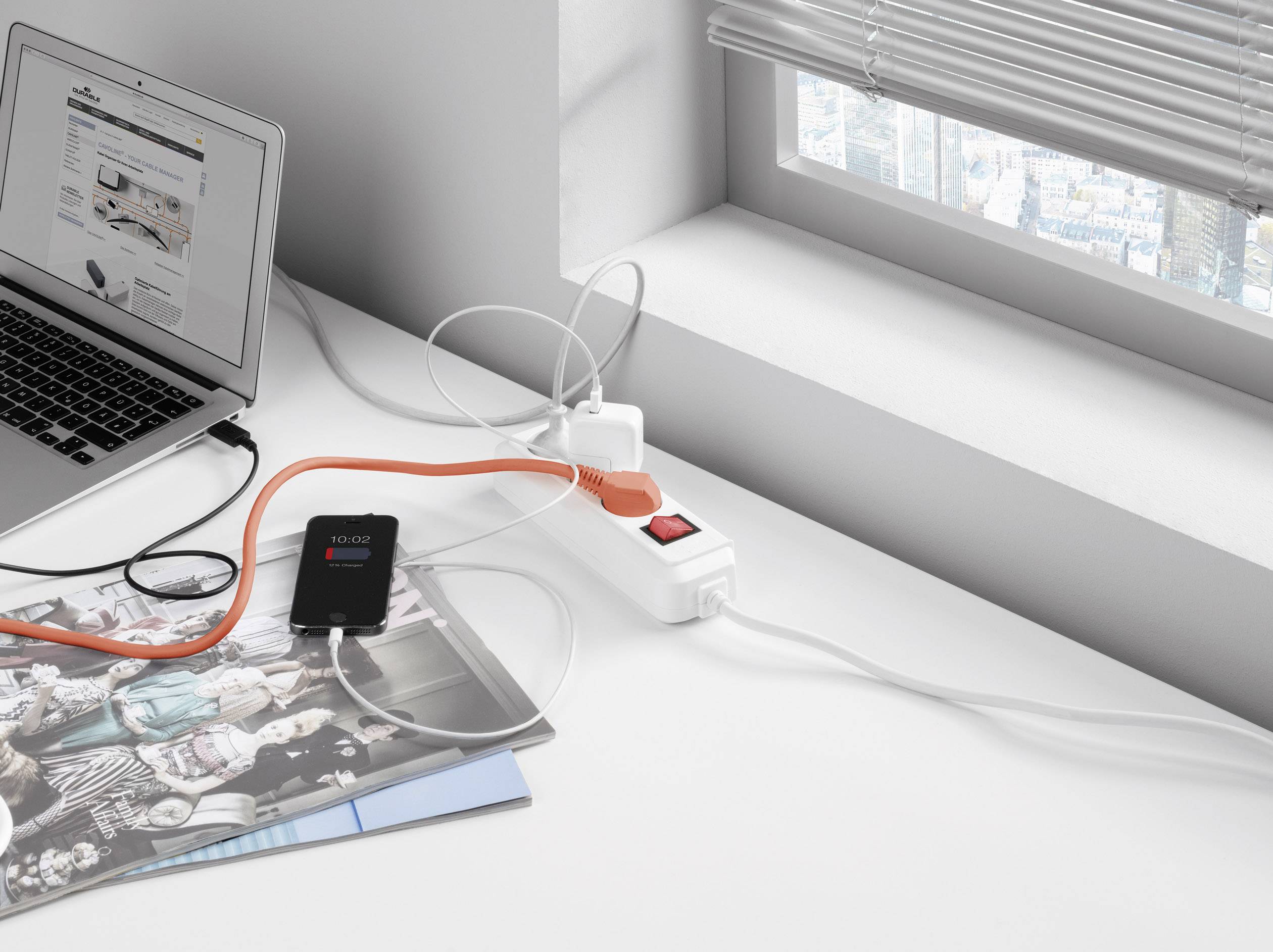 A laptop with an open webpage, connected to a multi-socket extension lead with several devices, on a desk by the window.