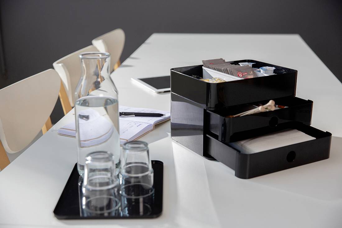 A modern conference table with a water jug, two glasses on a tray and an organiser with office supplies next to a notebook.